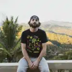 A person in a black t-shirt and cap sits on a ledge with a scenic mountain view in the background, creating the perfect spot for finding inspiration for new music.