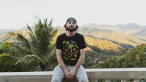 A person in a black t-shirt and cap sits on a ledge with a scenic mountain view in the background, creating the perfect spot for finding inspiration for new music.