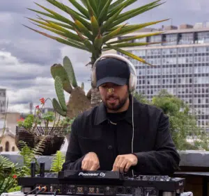 DJ wearing headphones, performing at an outdoor event. Plants and a cityscape are in the background.
