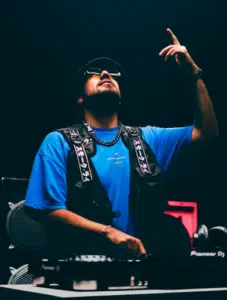 A DJ wearing sunglasses and a hat is performing on stage, pointing upward with one hand while using a turntable with the other.