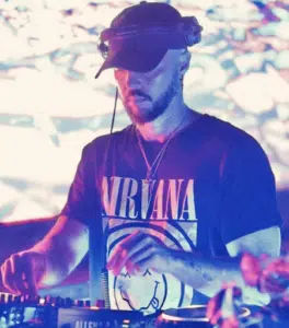 DJ wearing headphones and a Nirvana t-shirt, performing with DJ equipment in front of a colorful, abstract background.