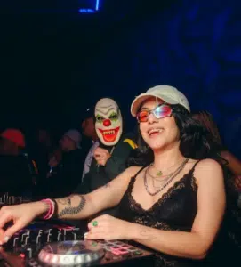 A person in a lace top and sunglasses smiles while DJing, with another person in a clown mask in the background.