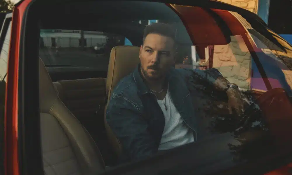 A man sits in a parked red car, gazing thoughtfully out the window as new music softly plays in the background.