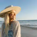 A person in overalls and a wide-brimmed hat smiles while standing on a beach, with the ocean harmonizing like new music in the background.