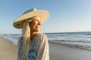 A person in overalls and a wide-brimmed hat smiles while standing on a beach, with the ocean harmonizing like new music in the background.