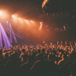 A large crowd stands with raised hands at a concert or live event, illuminated by vibrant purple and orange stage lights, their excitement tangible in the electric atmosphere.