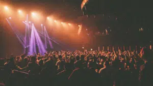 A large crowd stands with raised hands at a concert or live event, illuminated by vibrant purple and orange stage lights, their excitement tangible in the electric atmosphere.