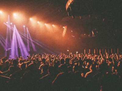 A large crowd stands with raised hands at a concert or live event, illuminated by vibrant purple and orange stage lights, their excitement tangible in the electric atmosphere.