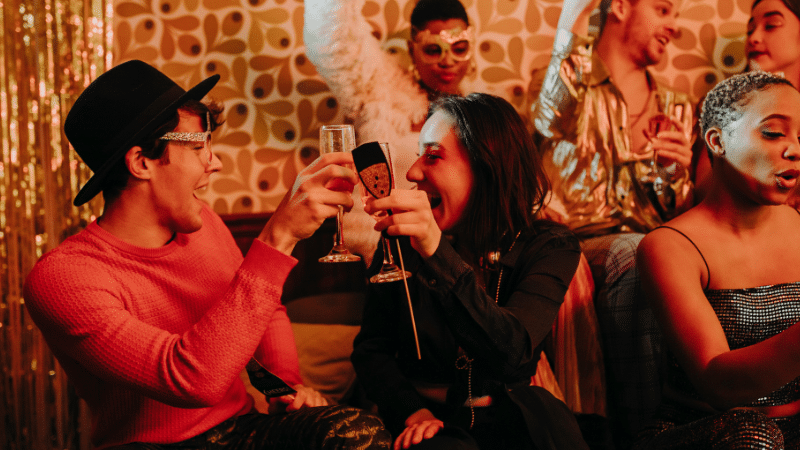 A lively group of people celebrating at a party, toasting with raised glasses. They're dressed in festive, colorful attire with some wearing trendy hats and masks against a retro patterned backdrop.