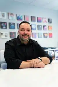 A man with a beard and black shirt sits at a white table, smiling as if reflecting on the year in review, with colorful framed pictures on the wall behind him.