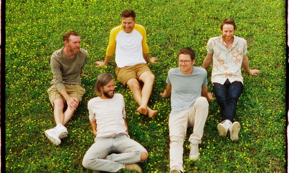 Five men sitting and laughing on a grassy field with scattered small yellow flowers, sharing the joy of discovering new music together.