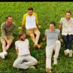 Five men sitting and laughing on a grassy field with scattered small yellow flowers, sharing the joy of discovering new music together.