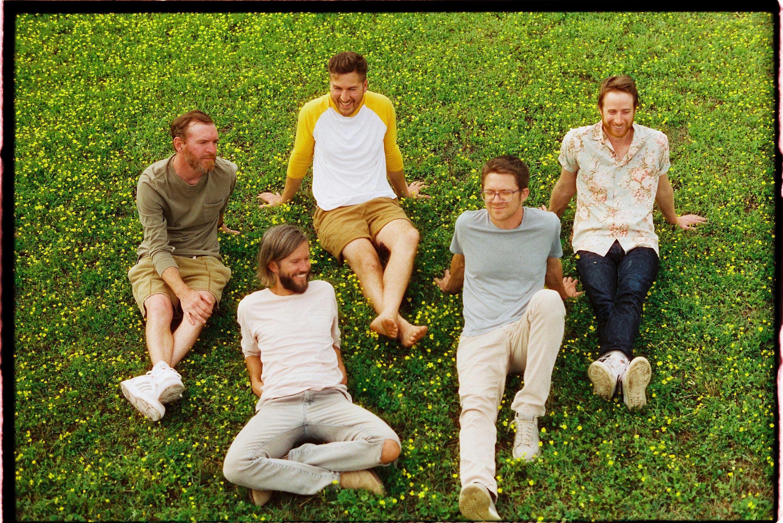 Five men sitting and laughing on a grassy field with scattered small yellow flowers, sharing the joy of discovering new music together.