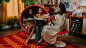 Three women in sparkly outfits sit on a red chair, exchanging gifts together in a retro-styled room with patterned wallpaper and a bold red carpet.