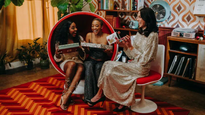 Three women in sparkly outfits sit on a red chair, exchanging gifts together in a retro-styled room with patterned wallpaper and a bold red carpet.