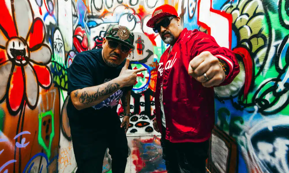 Two men pose in a graffiti-covered alley, one in a red jacket and cap, the other in a black shirt and cap, making gestures towards the camera as they announce their new music.