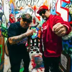 Two men pose in a graffiti-covered alley, one in a red jacket and cap, the other in a black shirt and cap, making gestures towards the camera as they announce their new music.