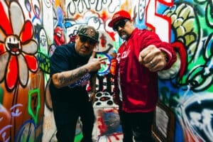 Two men pose in a graffiti-covered alley, one in a red jacket and cap, the other in a black shirt and cap, making gestures towards the camera as they announce their new music.