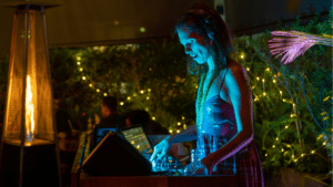 A woman in headphones operates DJ equipment at an outdoor event, illuminated by blue and yellow lights, with string lights and greenery in the background—an inspiring scene for those seeking grants for musicians.