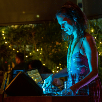 A woman in headphones operates DJ equipment at an outdoor event, illuminated by blue and yellow lights, with string lights and greenery in the background—an inspiring scene for those seeking grants for musicians.