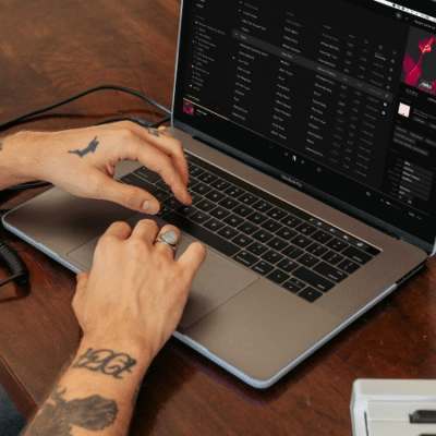 A person with tattoos uses a laptop displaying a music playlist, perhaps ready to claim your artist page. A coiled cable and a white electronic device are also on the wooden table.