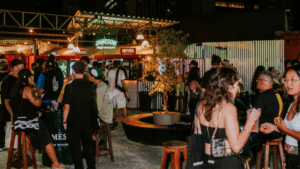 A group of people socialize at an outdoor bar at night, with drinks in hand and illuminated signs in the background, reminiscent of lively gatherings after music conferences.