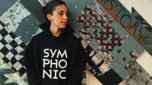 Person wearing a "SYMPHONIC" hoodie stands in front of a textured abstract wall with layered designs.