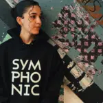 Person in a black hoodie with "SYMPHONIC" text, standing in front of an abstract wall art.