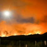 A wildfire burns intensely at night, illuminating the sky with orange flames and smoke, as a bright full moon offers its gentle support from above.