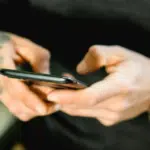 A person with tattooed hands holds a smartphone. They are wearing a black long-sleeve shirt and a large gold wristwatch.