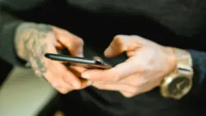 A person with tattooed hands holds a smartphone. They are wearing a black long-sleeve shirt and a large gold wristwatch.