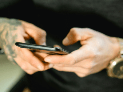 A person with tattooed hands holds a smartphone. They are wearing a black long-sleeve shirt and a large gold wristwatch.