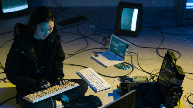 In a dim-lit room, a person in a dark jacket sits on the floor, engrossed in typing on a keyboard. They are surrounded by old monitors and tangled cables, with screens displaying blue and green hues—an intriguing scene of artificial streaming at its finest.