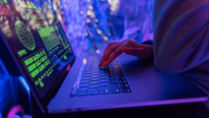 A person types on a laptop under vibrant green and pink neon lighting, streaming artificial data across the screen.