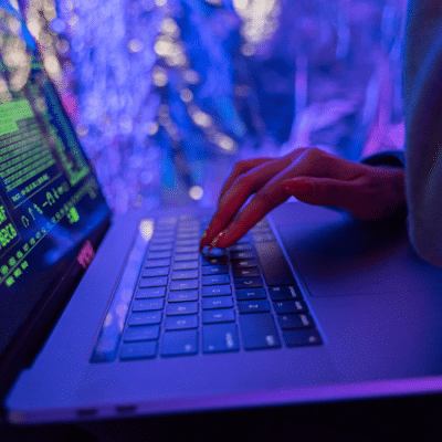 A person types on a laptop under vibrant green and pink neon lighting, streaming artificial data across the screen.