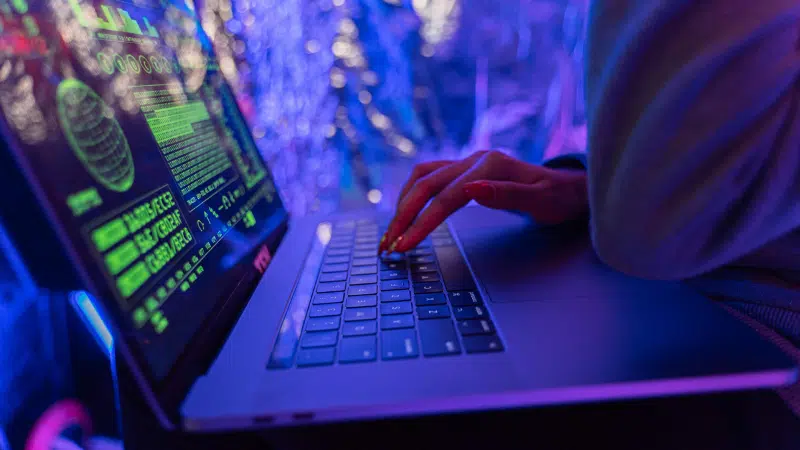 A person types on a laptop under vibrant green and pink neon lighting, streaming artificial data across the screen.