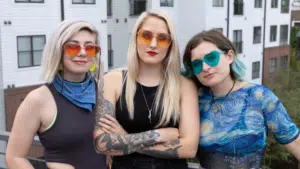 Three people with colorful sunglasses and casual attire stand outdoors in front of a building.