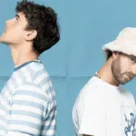 Two men stand back-to-back. One wears a striped shirt, looking up. The other wears a bucket hat and a graphic tee, looking forward against a blue background.