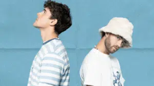 Two men stand back-to-back. One wears a striped shirt, looking up. The other wears a bucket hat and a graphic tee, looking forward against a blue background.