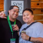 Two people are smiling inside a venue with wooden walls and plants, clearly enjoying a networking event. One person, wearing a hat and green lanyard, holds a drink as they stand before a sign marked "SYM.