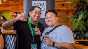 Two people are smiling inside a venue with wooden walls and plants, clearly enjoying a networking event. One person, wearing a hat and green lanyard, holds a drink as they stand before a sign marked "SYM.