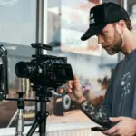 A man operates a professional camera on a tripod outside a shop, capturing scenes for a music video. He wears a cap and t-shirt, tattoos visible on his arms. Beside him, another person is partially visible.