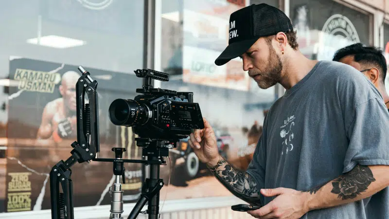 A man operates a professional camera on a tripod outside a shop, capturing scenes for a music video. He wears a cap and t-shirt, tattoos visible on his arms. Beside him, another person is partially visible.