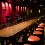 A dimly lit music venue in CDMX features a long bar with red-cushioned stools, wooden tables, and a stage adorned with musical instruments against a striking red curtain backdrop.