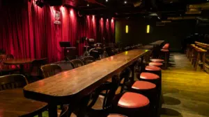 A dimly lit music venue in CDMX features a long bar with red-cushioned stools, wooden tables, and a stage adorned with musical instruments against a striking red curtain backdrop.