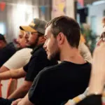 A group of people sit in a row at an indoor event. They appear attentive and engaged. Colorful banners hang above them.