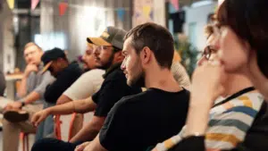 A group of people sit in a row at an indoor event. They appear attentive and engaged. Colorful banners hang above them.