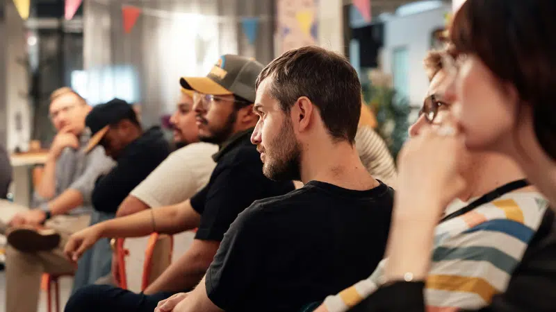 A group of people sit in a row at an indoor event. They appear attentive and engaged. Colorful banners hang above them.