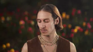 A person with medium-length hair and layered necklaces looks down, standing in front of a blurred background with red and yellow flowers.