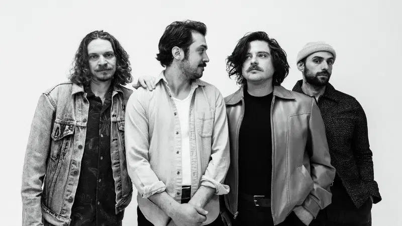 Four men stand together against a plain background. They appear to be in casual attire, with one wearing a beanie. The image is black and white.